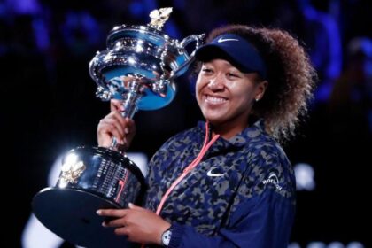 Naomi Osaka holds the Australian Open trophy in 2021. She is wearing a blue and grey tracksuit jacket with a pink zip, has a blue visor on her head and is smiling broadly.